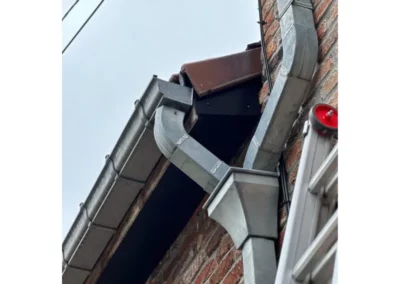 Raccordement de la descente d'eau pluviale en zinc à partir de la gouttière installée sur la toiture rénovée à Roux. Ce système assure un écoulement efficace de l'eau de pluie pour protéger les fondations de la maison.