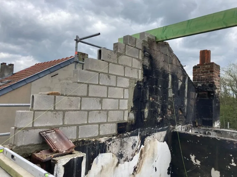 Mur pignon en cours de reconstruction, avec des blocs de béton servant à renforcer la structure. Également une poutre verte destinée à soutenir la nouvelle charpente de la toiture. Les travaux sont réalisés par TS Rénovation pour stabiliser et rénover la maison à Roux.